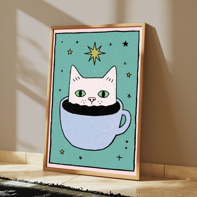 May include: A framed wall art print featuring a light pink cat with green eyes sitting inside a light blue coffee cup. The background is a textured teal color decorated with small black stars and a single yellow star above the cat's head.