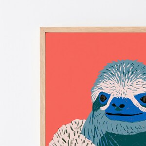 May include: A framed wall art print featuring a stylized illustration of a sloth with a blue face and textured fur in shades of blue, white, and dark green, set against a solid coral-colored background within a light-toned wooden frame.