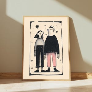 May include: A framed wall art print featuring a black and white illustration of two figures standing side by side. The figure on the left wears a grey top and black trousers, while the figure on the right wears a black top and pink trousers, all within a rectangular border with small star and crescent moon accents.