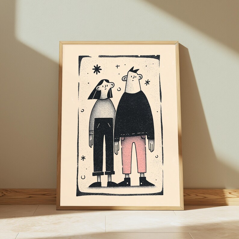 May include: A framed wall art print featuring a black and white illustration of two figures standing side by side. The figure on the left wears a grey top and black trousers, while the figure on the right wears a black top and pink trousers, all within a rectangular border with small star and crescent moon accents.