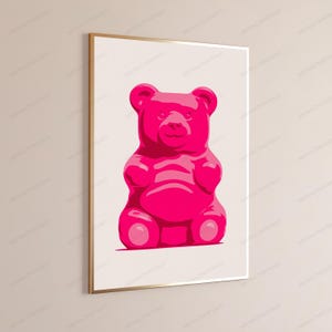 May include: A framed wall art print featuring a vibrant hot pink gummy bear illustration centered on a light cream background. The artwork is enclosed in a thin metallic gold frame, showcasing a clean, graphic design with bold, saturated colors.