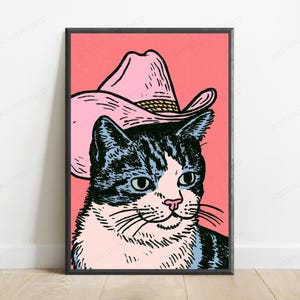May include: A framed wall art print featuring a graphic illustration of a cat wearing a pink cowboy hat. The cat has black and white fur, green eyes, and a pink nose. The background is a solid coral pink color, and the print is encased in a black frame.
