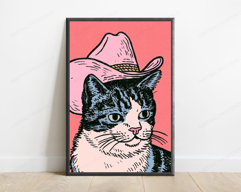 May include: A framed wall art print featuring a graphic illustration of a cat wearing a pink cowboy hat. The cat has black and white fur, green eyes, and a pink nose. The background is a solid coral pink color, and the print is encased in a black frame.