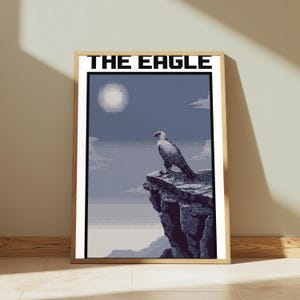 May include: A framed pixel art print titled THE EAGLE, featuring a white and dark grey eagle perched on a jagged cliff edge against a blue sky with a pale sun and clouds. The artwork uses a blocky, digital aesthetic with a muted colour palette.
