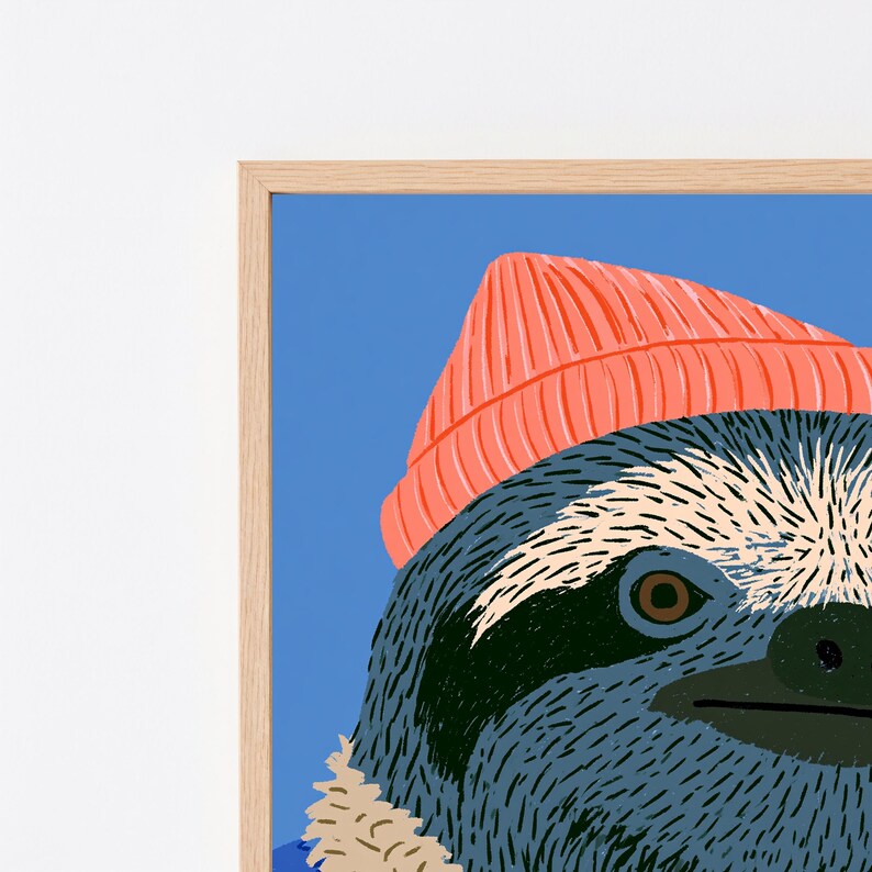May include: A framed art print featuring a stylized illustration of a sloth wearing a bright coral-colored beanie. The sloth has dark gray fur with cream-colored accents around the face and eyes, set against a solid medium blue background.