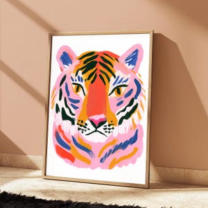 May include: A framed art print featuring a stylized tiger head illustration. The tiger is rendered with bold, expressive brushstrokes in vibrant shades of pink, orange, deep blue, and black against a clean white background within a light wood frame.