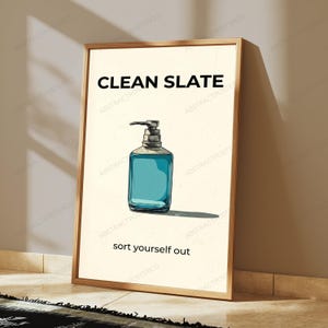 May include: A rectangular framed wall art print featuring a cream-colored background with a central illustration of a blue liquid soap dispenser. The text CLEAN SLATE appears in black at the top, and the text sort yourself out is at the bottom.