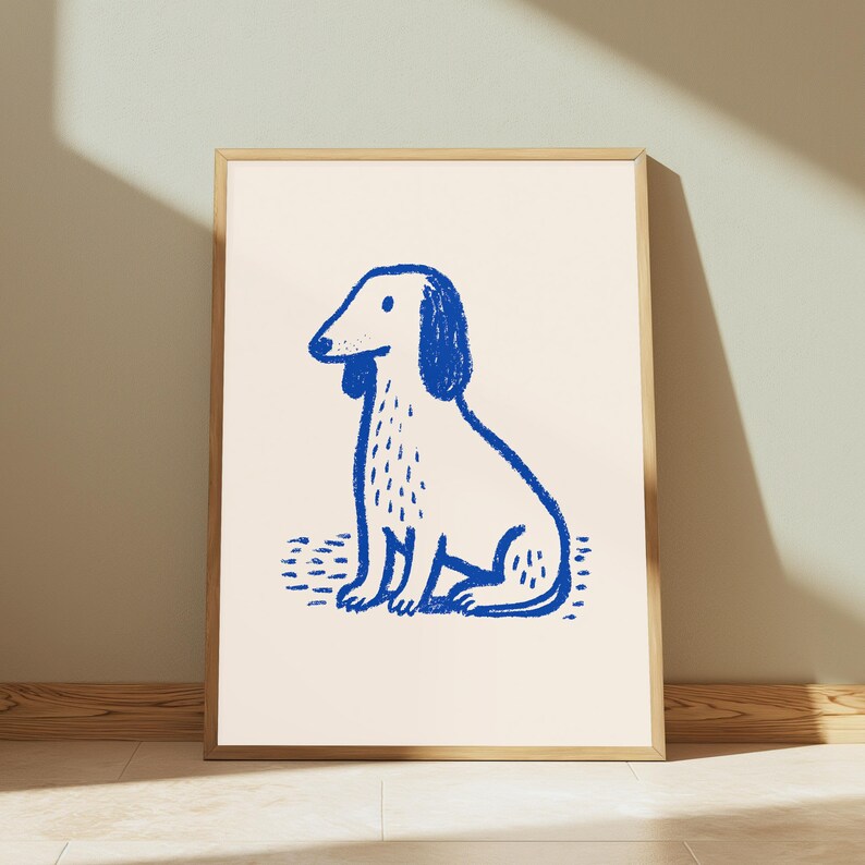 K&ouml;nnte beinhalten: Ein gerahmter Kunstdruck mit einer blauen Strichzeichnung eines sitzenden Hundes auf cremefarbenem Hintergrund. Die Illustration verwendet einen strukturierten, kreideartigen Strich, um die Hundeform darzustellen, die mittig im Holzrahmen platziert ist.