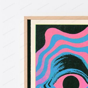 May include: A framed wall art print featuring a graphic design with wavy, concentric bands of bright pink and light blue against a dark black background. The artwork is enclosed in a light-colored wooden frame with a visible grain texture.