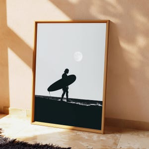 May include: A framed vertical wall art print featuring a black and white silhouette of a person carrying a surfboard along the shoreline. The scene includes a bright full moon in the sky above the dark ocean waves and the sandy beach area.