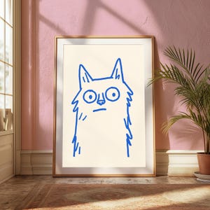 May include: A framed wall art print featuring a simple, hand-drawn blue line illustration of a cat with wide, circular eyes and pointed ears centered on a cream-colored background. The artwork is enclosed in a thin, metallic gold-colored frame.