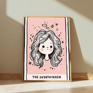 May include: A framed rectangular wall art print featuring a light pink background with a black line drawing of a person with wavy hair. Above the head are small fish and star icons. The text THE OVERTHINKER is printed in black at the bottom.