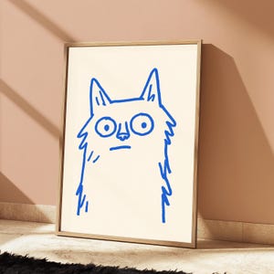 May include: A framed wall art print featuring a simple, hand-drawn illustration of a cat in bright blue ink on a cream-colored background. The cat has large, wide eyes, pointed ears, and a straight line for a mouth, rendered in a bold, sketchy style.