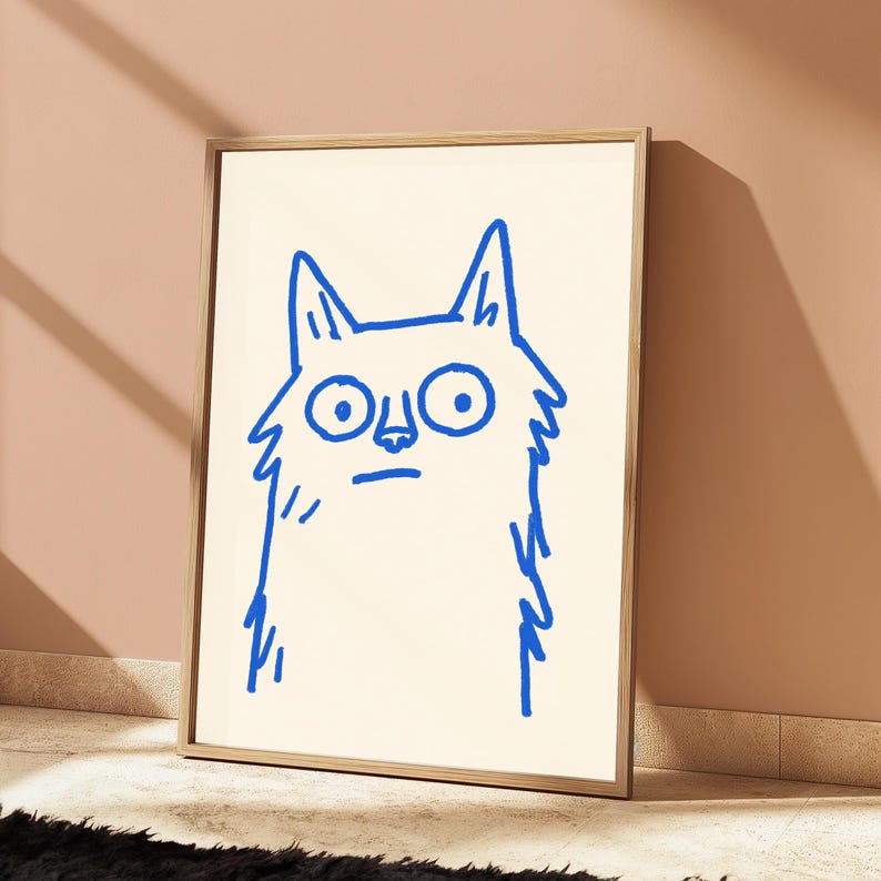 May include: A framed wall art print featuring a simple, hand-drawn illustration of a cat in bright blue ink on a cream-colored background. The cat has large, wide eyes, pointed ears, and a straight line for a mouth, rendered in a bold, sketchy style.