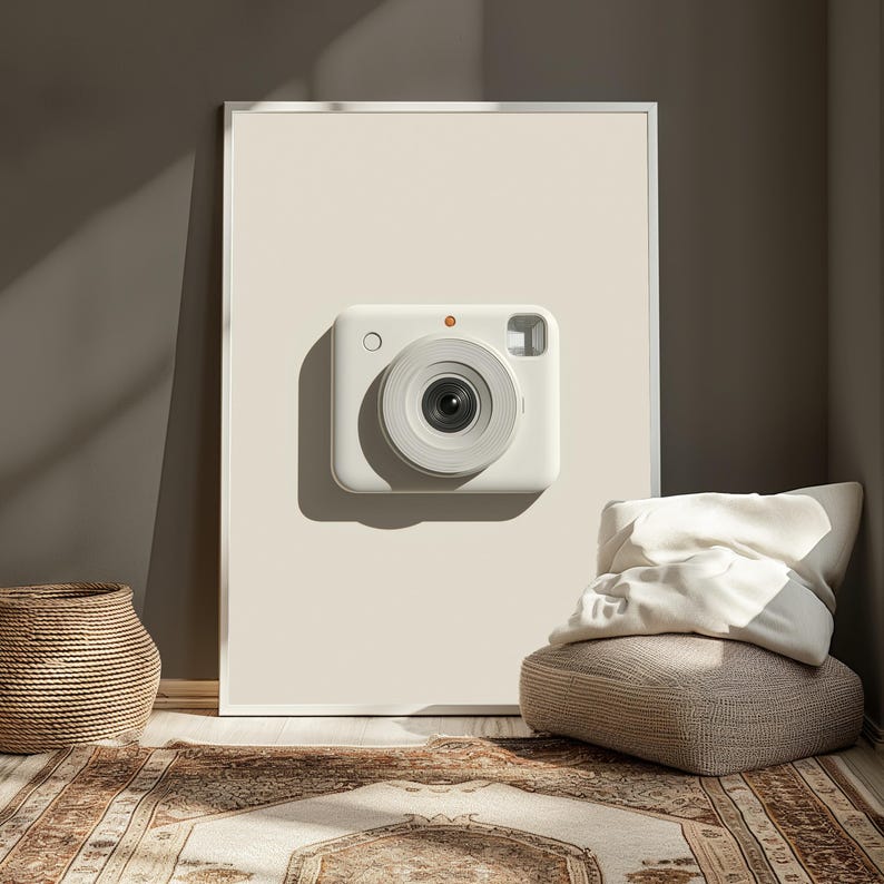 May include: A rectangular wall art print featuring a centered, minimalist illustration of a white instant camera with a prominent circular lens, a small orange dot above the lens, and a square viewfinder, all set against a solid cream background.