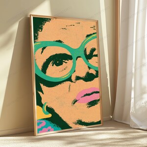 May include: A framed vertical art print featuring a stylized, high-contrast portrait of a person wearing large, bright green glasses. The image uses a peach-toned background with dark green shadows, a vibrant pink mouth, and a yellow earring detail.