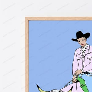 May include: A framed wall art print featuring a stylized illustration of a figure in a black cowboy hat, a light pink western shirt with a black and white polka dot neckerchief, and blue jeans, holding the reins of a longhorn steer against a blue background.