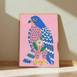 May include: A framed art print featuring a stylized blue falcon on a solid pink background. The bird has a patterned chest with blue dots and a central white flower with green leaves. The falcon has yellow talons and a blue and white wing design.