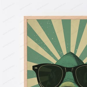 May include: A framed wall art print featuring a green creature wearing black sunglasses. The background displays a sunburst pattern with alternating light beige and muted green rays, accented by small white dots across the entire surface.