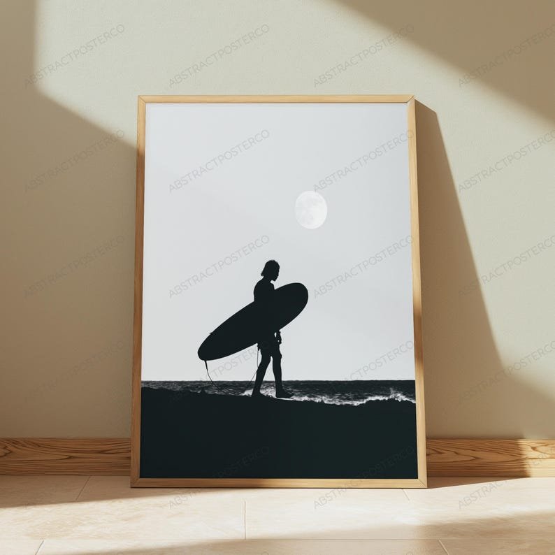 May include: A framed black and white wall art print featuring a silhouette of a person carrying a surfboard along a shoreline. The scene depicts a dark beach, a calm ocean, and a bright circular moon in the sky, all enclosed in a light wood frame.