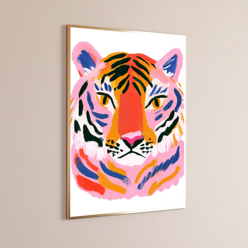 May include: A framed wall art print featuring a stylized tiger head illustration. The tiger is rendered in vibrant shades of pink, orange, and deep blue, with bold black stripes and bright yellow eyes, set against a clean white background.