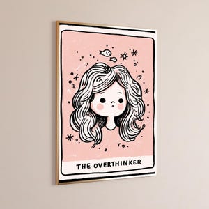 May include: A rectangular wall art print featuring a pale pink background with a black-lined illustration of a person with wavy hair and rosy cheeks. Small fish and star icons surround the head. The text THE OVERTHINKER is printed at the bottom.
