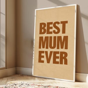 May include: A framed print with the words "BEST MUM EVER" in brown block letters on a tan background. The print is in a white frame and leans against a wall. The room has a window and a patterned rug.