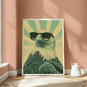 May include: A rectangular wall art print featuring a stylized illustration of a bald eagle wearing black sunglasses. The eagle is centered against a background of alternating light yellow and muted green sunburst rays with a dotted texture pattern.