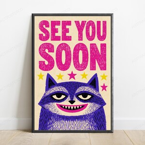 May include: A framed rectangular poster with a cream background featuring the text SEE YOU SOON in large, distressed pink letters. Below the text is a stylized purple raccoon with a wide grin, sharp teeth, and a row of yellow and pink stars.