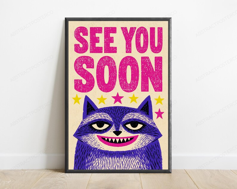 May include: A framed rectangular poster with a cream background featuring the text SEE YOU SOON in large, distressed pink letters. Below the text is a stylized purple raccoon with a wide grin, sharp teeth, and a row of yellow and pink stars.
