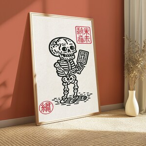 May include: A framed wall art print featuring a black ink illustration of a skeleton holding a small rectangular tablet. The skeleton is depicted in a cartoon style on a white background with red stamp-like characters in the corners.