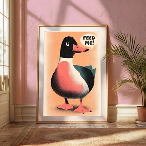 May include: A framed wall art print featuring a stylised illustration of a duck with a black head, a pink chest, and dark wings against a light orange background. A white speech bubble above the duck contains the black text FEED ME!