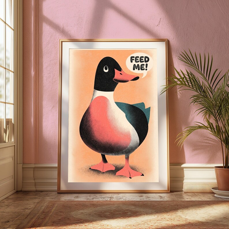 May include: A framed wall art print featuring a stylised illustration of a duck with a black head, a pink chest, and dark wings against a light orange background. A white speech bubble above the duck contains the black text FEED ME!