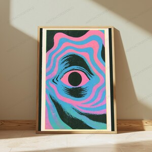 May include: A rectangular wall art print in a light wood frame featuring a central black pupil surrounded by concentric, wavy bands of bright pink and cyan blue. The design includes dark, textured brushstroke patterns that radiate outward.