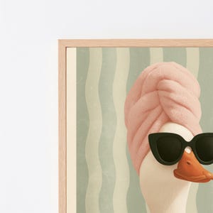 Puede incluir: Una l&aacute;mina de arte mural enmarcada que muestra un ganso blanco con una toalla rosa envuelta alrededor de la cabeza y gafas de sol negras de ojo de gato. El fondo presenta rayas verticales onduladas en tonos verde claro y crema, con marco de madera clara.