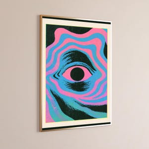 May include: A rectangular wall art print in a thin gold frame featuring a central black pupil surrounded by concentric, wavy lines in alternating shades of bright pink and cyan against a dark, textured black background with a white border.