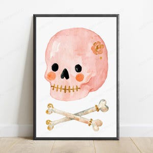 May include: A framed wall art print featuring a watercolor illustration of a light pink skull with black eyes, orange cheek spots, and a gold stitched mouth. A small gold flower sits on the side of the skull, above two crossed beige bones.