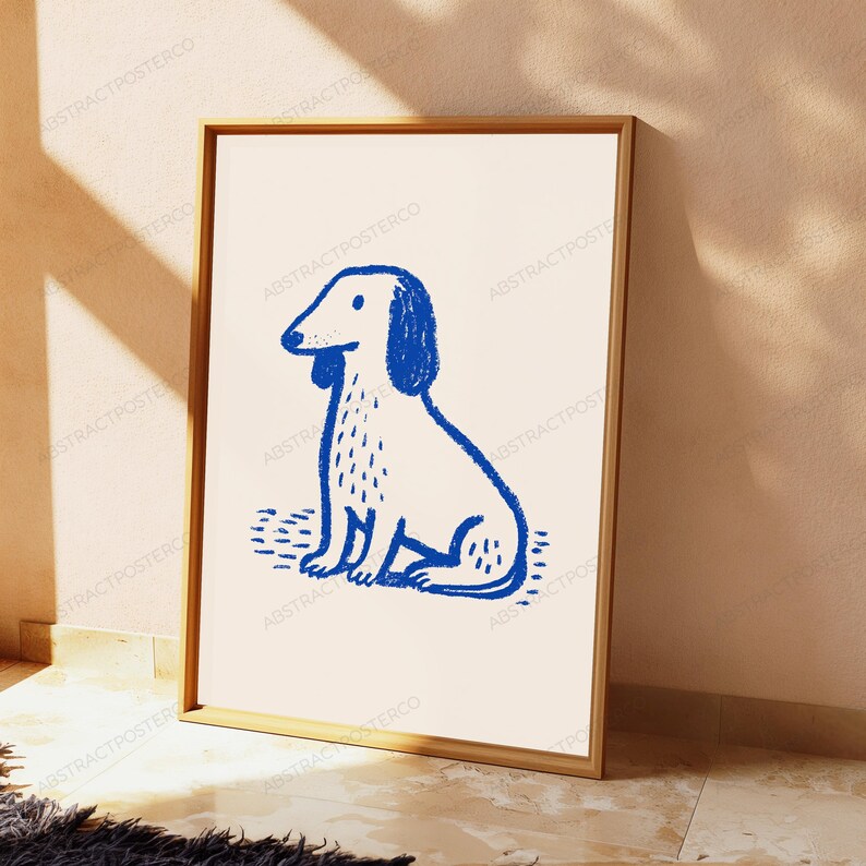 May include: A framed wall art print featuring a blue line drawing of a sitting dog on a cream background. The dog illustration has a textured, crayon-like appearance with simple lines defining its ears, body, and tail. The frame is light wood.