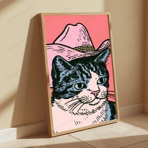 May include: A framed wall art print featuring a black and white cat wearing a pink cowboy hat against a solid pink background. The illustration uses bold black ink lines to define the cat's facial features and the texture of the hat.