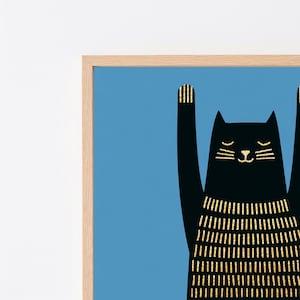 Op de afbeelding: Een ingelijste kunstprint met het silhouet van een zwarte kat tegen een effen blauwe achtergrond. De kat heeft de voorpoten omhoog en is versierd met verticale gouden glitterstrepen op het lichaam, de oren en de poten, in een lichthouten lijst.