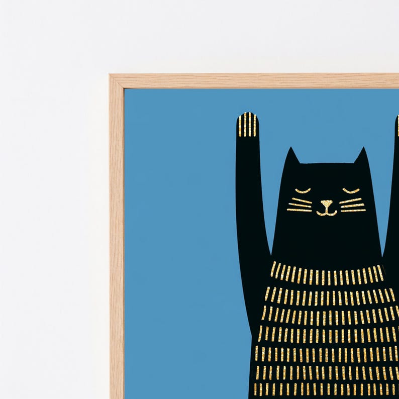 Op de afbeelding: Een ingelijste kunstprint met het silhouet van een zwarte kat tegen een effen blauwe achtergrond. De kat heeft de voorpoten omhoog en is versierd met verticale gouden glitterstrepen op het lichaam, de oren en de poten, in een lichthouten lijst.