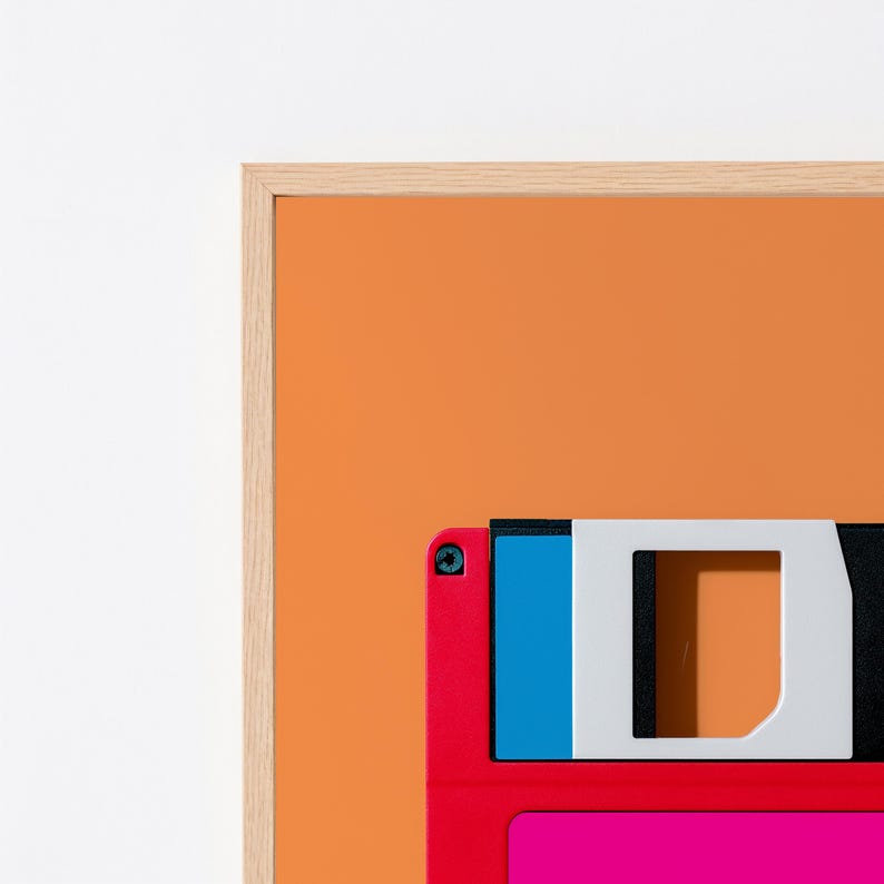 May include: A framed wall art print featuring a vibrant orange background with a partial view of a retro floppy disk. The disk displays a bright red frame, a blue accent section, and a white sliding metal shutter, all encased in a light wood frame.