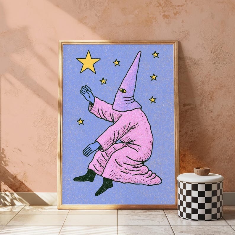 May include: A framed wall art print featuring a figure in a pink hooded robe with a single eye on the hood, reaching toward yellow stars against a textured light blue background. The figure has blue hands and black feet, depicted in a stylized illustration.