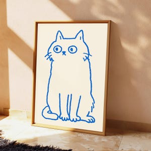 May include: A framed wall art print featuring a simple, hand-drawn blue outline of a cat with wide eyes and a small nose on a cream-colored background. The cat is depicted in a seated position with a textured, crayon-like stroke style.