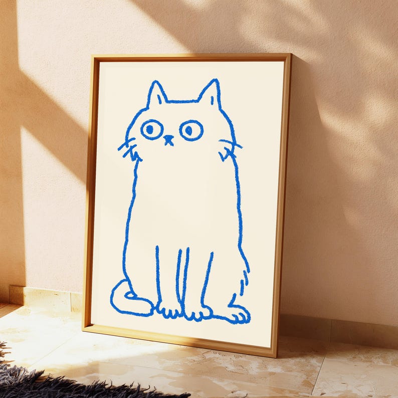 May include: A framed wall art print featuring a simple, hand-drawn blue outline of a cat with wide eyes and a small nose on a cream-colored background. The cat is depicted in a seated position with a textured, crayon-like stroke style.