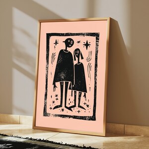 May include: A framed wall art print featuring a black ink illustration on a light peach background. The artwork depicts two stylized figures standing side by side within a textured rectangular border, surrounded by small star and curved line motifs.