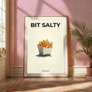 May include: A framed wall art print featuring a cream-colored background with a central illustration of golden-brown french fries in a white container. The text BIT SALTY appears in bold black letters above, with the word standard printed below.