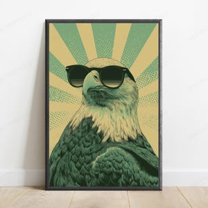May include: A framed wall art print featuring a stylized illustration of a bald eagle wearing black sunglasses. The eagle is set against a background of alternating light green and tan sunburst rays with a subtle dotted texture throughout the design.