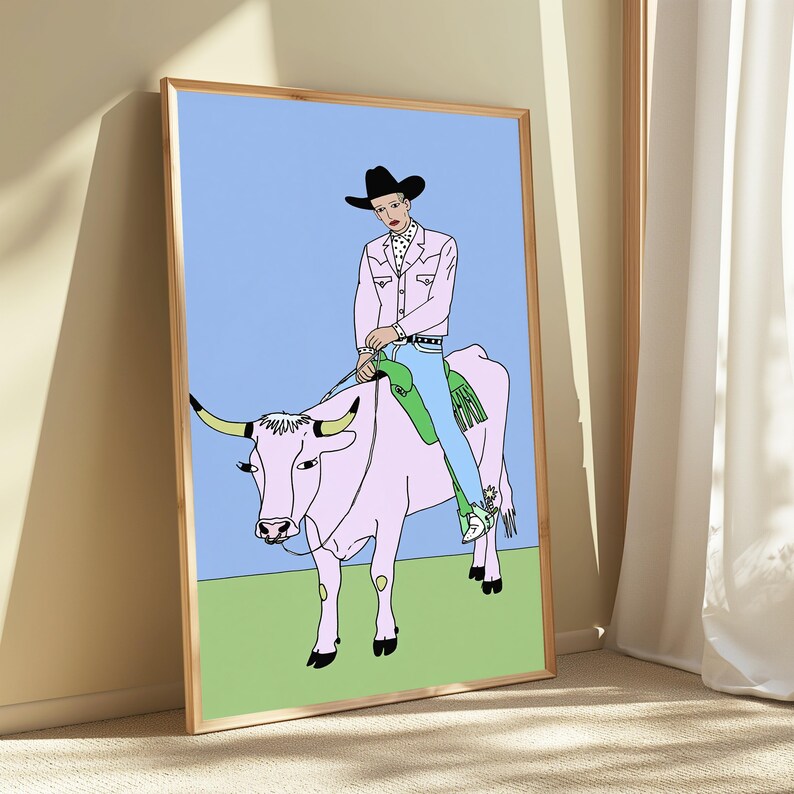 May include: A framed art print featuring a line-art illustration of a person in a black cowboy hat, pink jacket, and blue jeans riding a large pink bull. The bull has long horns and a nose ring, set against a light blue sky and green ground.