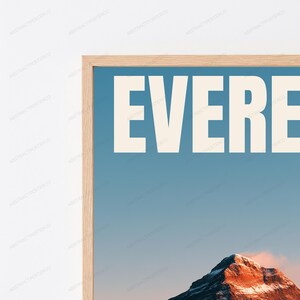 Op de afbeelding: Een ingelijste kunstdruk met een lichtblauwe lucht op de achtergrond en de grote witte tekst EVEREST bovenaan. Onder de tekst is een besneeuwde bergtop te zien die wordt verlicht door warm, oranje zonlicht tegen de blauwe lucht.